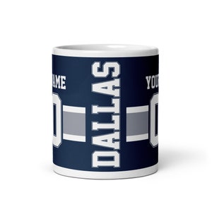 K&ouml;nnte beinhalten: Wei&szlig;e Keramiktasse mit blauem und grauem Design. Die Tasse zeigt das Wort "DALLAS" in wei&szlig;en Buchstaben und ein graues und wei&szlig;es Streifenmuster. Die Tasse ist mit dem Text "NAME" und "YOUR NUMBER" in wei&szlig;en Buchstaben personalisiert.