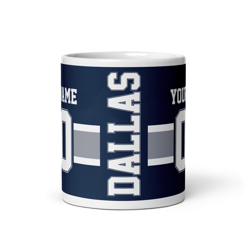 K&ouml;nnte beinhalten: Wei&szlig;e Keramiktasse mit blauem und grauem Design. Die Tasse zeigt das Wort "DALLAS" in wei&szlig;en Buchstaben und ein graues und wei&szlig;es Streifenmuster. Die Tasse ist mit dem Text "NAME" und "YOUR NUMBER" in wei&szlig;en Buchstaben personalisiert.