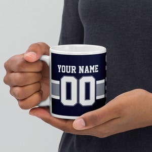 K&ouml;nnte beinhalten: Wei&szlig;e Keramiktasse mit einem marineblauen und silbernen Design. Die Tasse zeigt eine wei&szlig;e Nummer "00" und den Text "YOUR NAME".