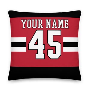 Könnte beinhalten: Ein rotes, schwarzes und weißes Kissen mit einem Trikot-Design. Das Kissen zeigt den Text "YOUR NAME" und die Nummer "45" in Weiß.