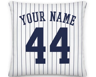 May include: A square throw pillow featuring a white background with vertical navy blue pinstripes. The text "YOUR NAME" is printed in a navy blue athletic font above the large navy blue number "44" centered on the front of the pillow.
