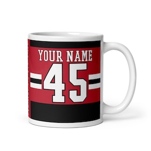 May include: White ceramic mug with a red, black, and white design. The design features the text "YOUR NAME" and the number "45" in a white jersey-style font.