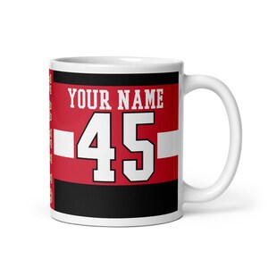 May include: White ceramic mug with a red, black, and white design. The design features the text "YOUR NAME" and the number "45" in white.