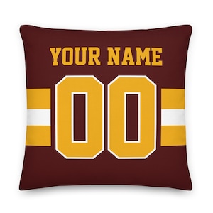 Puede incluir: Una almohada de color marrón con un diseño de camiseta de fútbol dorada y blanca. La almohada presenta el texto "YOUR NAME" y el número "00".