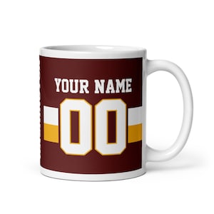 May include: White ceramic mug with a maroon background and a white and gold football jersey design. The jersey has the text "YOUR NAME" and the number "00" on it.
