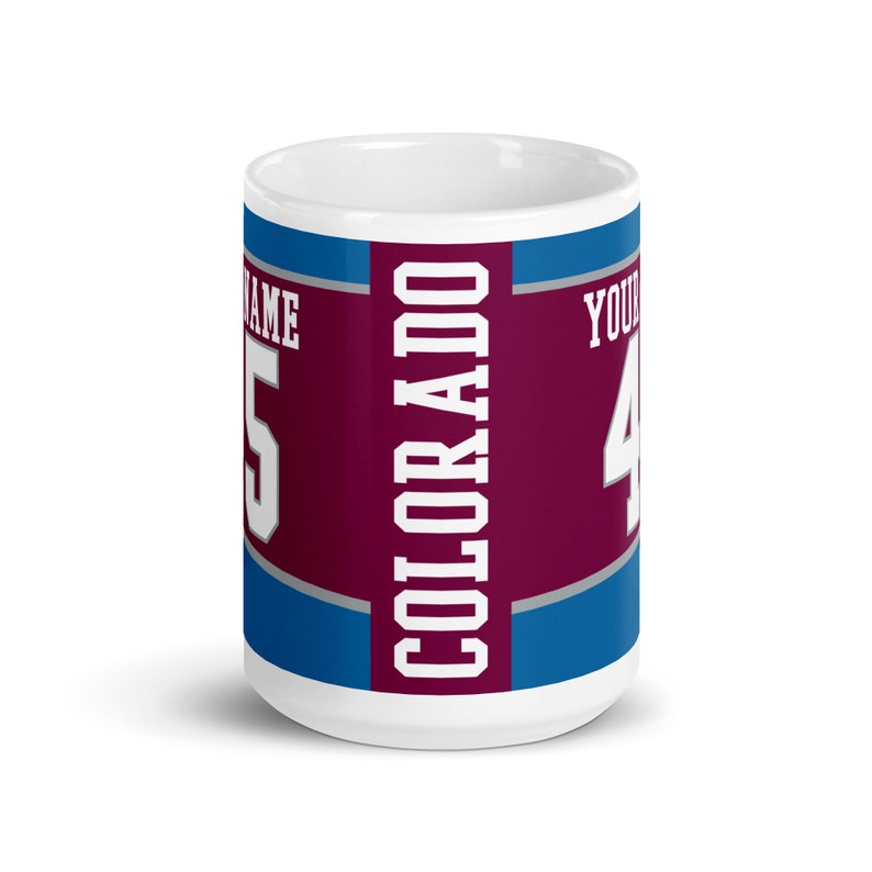 May include: White ceramic coffee mug with a burgundy, blue, and gray design. The word "COLORADO" is printed vertically in white on the burgundy stripe. The number "5" is printed in gray on the left side of the mug and the number "4" is printed in gray on the right side of the mug.
