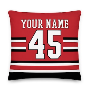 Könnte beinhalten: Ein rotes und schwarzes Kissen mit einem weißen Trikotdesign. Das Trikot trägt den Text "YOUR NAME" und die Nummer "45".