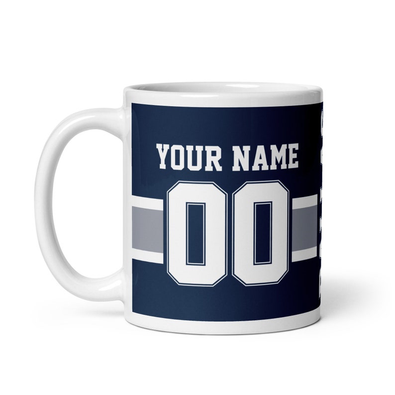K&ouml;nnte beinhalten: Wei&szlig;e Keramiktasse mit blauem und grauem Design. Die Tasse zeigt eine wei&szlig;e Nummer "00" und den Text "YOUR NAME".