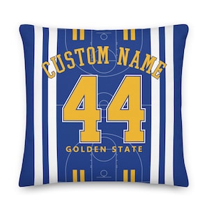 Könnte beinhalten: Ein quadratisches Kissen mit blau-weißem Streifenmuster. Es zeigt ein Basketballfeld-Design mit dem Text "CUSTOM NAME" und der Zahl "44" in Gold. Die Worte "GOLDEN STATE" sind ebenfalls sichtbar.