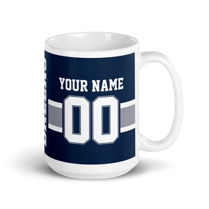 K&ouml;nnte beinhalten: Wei&szlig;e Keramiktasse mit blauem und grauem Design. Die Tasse zeigt eine wei&szlig;e Nummer "00" und den Text "YOUR NAME".