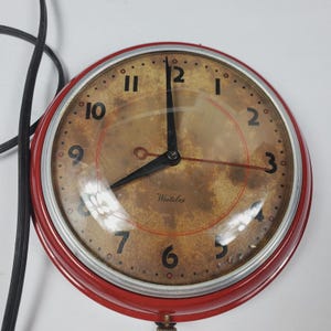 May include: A vintage round wall clock with a red metal frame. The clock face is a mottled brown with black numerals and hands. The clock reads "Westclox" at the bottom. A black power cord extends from the top left.