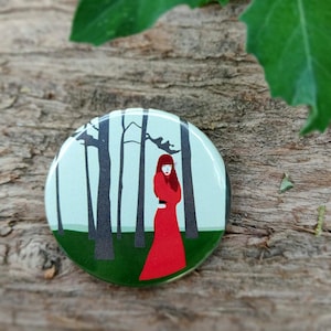 Puede incluir: Un botón redondo con una mujer con un vestido rojo de pie en un bosque. Los árboles son negros y el fondo es verde claro.