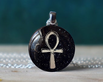 May include: A silver chain necklace with a black and silver pendant featuring an Ankh symbol on a black background with silver glitter.