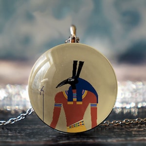 Op de afbeelding: Een zilveren ketting met een ronde glazen hanger met een kleurrijke illustratie van de Egyptische god Anubis. De hanger heeft een beige achtergrond met een zwart en blauw beeld van Anubis.