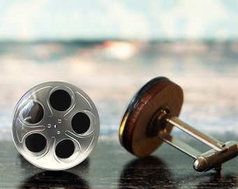May include: A pair of silver cufflinks featuring a film reel design. The cufflinks are made of metal and have a silver finish. The film reel is black and has a silver rim.