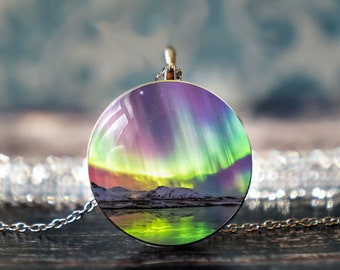 May include: A round silver pendant necklace with a colorful image of the aurora borealis reflected in a body of water. The image is set against a backdrop of snow-capped mountains.