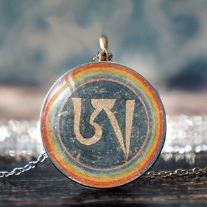 May include: A silver chain necklace with a round pendant featuring a rainbow border and a blue background with a white symbol in the center. The symbol is a stylized "Om" in Sanskrit.