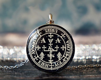 May include: A round, black pendant with a silver chain. The pendant features white text and symbols arranged in a circular pattern. The text includes the name "URIEL" and other words. The pendant has a silver bail.