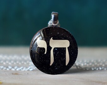 May include: A silver chain necklace with a round pendant featuring a white Hebrew letter "Chai" on a black background with silver glitter.