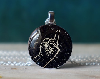 May include: A silver necklace with a round black pendant featuring a white silhouette of a hand giving the 'rock on' hand gesture against a black background with white glitter.