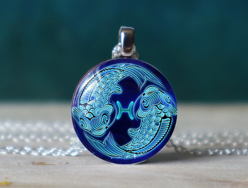 Puede incluir: Un collar de cadena de plata con un colgante redondo que presenta un s&iacute;mbolo del zod&iacute;aco de Piscis azul y verde. El colgante representa dos peces entrelazados con el s&iacute;mbolo de Piscis en el centro.