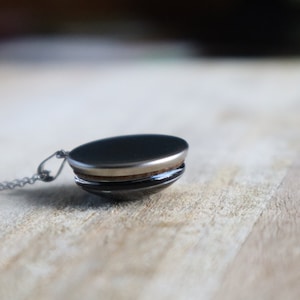 Puede incluir: Un collar de cadena de plata con un colgante redondo negro. El colgante tiene un borde plateado y una banda de madera alrededor del medio.