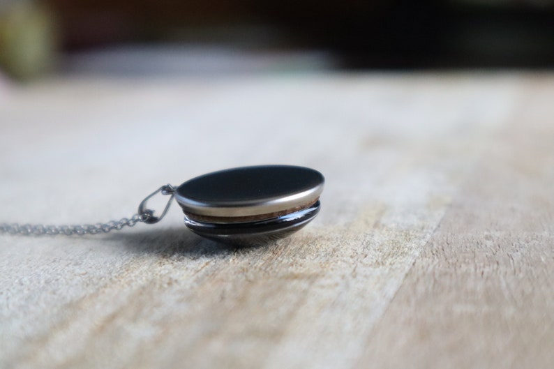 Puede incluir: Un collar de cadena de plata con un colgante redondo negro. El colgante tiene un borde plateado y una banda de madera alrededor del medio.