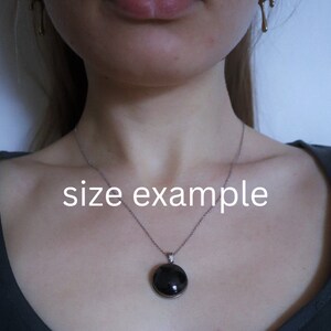 Puede incluir: Un collar de cadena de plata con un colgante redondo negro. El colgante tiene un di&aacute;metro de aproximadamente 2,5 cm. El collar lo lleva una persona, pero no se ve el rostro de la persona. El texto "size example" est&aacute; escrito en la imagen.