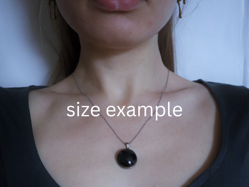 Puede incluir: Un collar de cadena de plata con un colgante redondo negro. El colgante tiene un di&aacute;metro de aproximadamente 2,5 cm. El collar lo lleva una persona, pero no se ve el rostro de la persona. El texto "size example" est&aacute; escrito en la imagen.