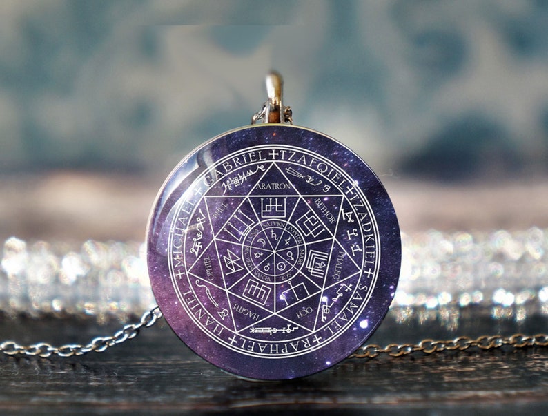 May include: A silver necklace with a round pendant featuring a white circle with a star of David and the names of the archangels in a circle around it. The pendant has a galaxy-like background.