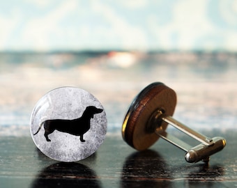 Puede incluir: Un par de gemelos con un conector de metal plateado. Un gemelo presenta una silueta negra de un dachshund sobre un fondo gris y blanco. El otro gemelo es de madera.