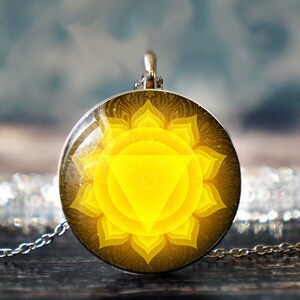 Puede incluir: Un collar de cadena de plata con un colgante redondo que presenta un mandala amarillo con un triángulo en el centro. El colgante está rodeado por un diseño negro y dorado.