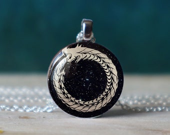 May include: A silver necklace with a round pendant featuring a white snake design on a black background with a starry pattern.