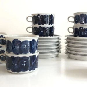 May include: A collection of white ceramic cups, saucers, and a lidded bowl, all adorned with a dark blue floral pattern. The cups have handles and are stacked, alongside stacks of saucers. The set is arranged on a white surface.