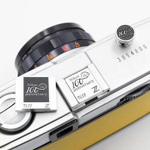 以下が含まれることがあります： 黒いレンズと黄色の革のトリムが付いた、銀色のニコンカメラのクローズアップ。 カメラには、いくつかの銀色のプレートと、銀色のボタンに「Nikon 100th anniversary」の文字が記されています。 シリアル番号「3054800」が表示されています。