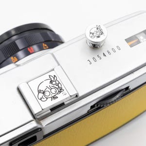 以下が含まれることがあります： 黒いレンズと黄色の革製グリップが付いたシルバーのフィルムカメラ。 カメラには、小さな丸いボタンと長方形のプレートがあり、それぞれに白黒の漫画のイラストが描かれています。 番号「3054800」がカメラ本体に印刷されています。