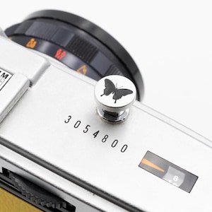 以下が含まれることがあります： 黒いレンズと蝶のデザインのボタンが付いた、銀色のカメラ本体のクローズアップ。ボタンは銀色で、黒い蝶が付いています。カメラには「3054800」の数字が印刷されています。