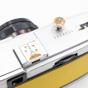 以下が含まれることがあります： 黄色の革製グリップが付いた、シルバーのヴィンテージカメラ。 カメラには、緑とオレンジのデザインの装飾ボタンと、フラッシュアタッチメントの小さなイラストがあります。 シリアル番号3054800が表示されています。