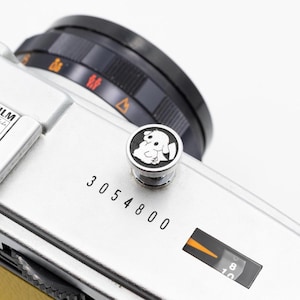 以下が含まれることがあります： 黒いレンズと漫画の犬のデザインの装飾ボタンが付いた、銀色のカメラのクローズアップ。 カメラ本体には「3054800」の数字と黒とオレンジのダイヤルが表示されています。