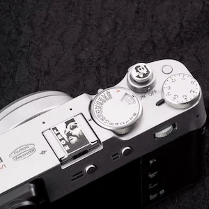 以下が含まれることがあります： 複数のダイヤルとボタンを備えた、シルバーとブラックのデジタルカメラのクローズアップ。 カメラには数字のダイヤルと人物の小さな画像があります。 「FUJINON LENS SYSTEM」の文字も表示されています。