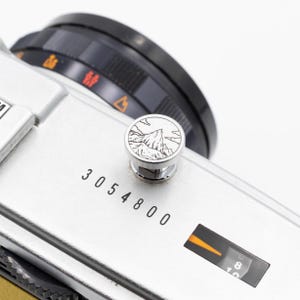 以下が含まれることがあります： 黒いレンズと山岳デザインが施されたシルバーのカメラのクローズアップ。カメラの本体には「3054800」の数字が印刷されています。レンズにはオレンジと白のマーキングがあります。