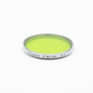 May include: A green Kodak filter with a silver rim. The filter is labeled "Kodak F III/32 2x".