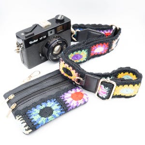 Puede incluir: Una cámara negra con una correa de cuero negro y una correa de cámara de crochet de colores. La cámara tiene el texto "Rollei XF 35" en la parte delantera. También se incluye una cartera de cuero negro con cremallera y un diseño de crochet de colores.