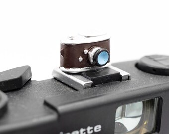 May include: A miniature brown and silver camera charm with a blue lens is mounted on a black camera body. The camera body has the text "beirette" and "gen 2" printed on it. The background is white.