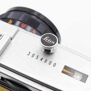 以下が含まれることがあります： 黒いレンズと黒とシルバーのライカのロゴボタンが付いた、シルバーのライカカメラ本体のクローズアップ。シリアル番号3054800がカメラ本体に印刷されています。カメラの設定が表示されています。