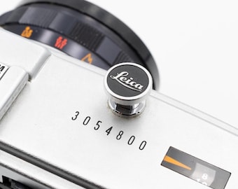 May include: Close-up of a silver Leica camera body with a black lens and a black and silver Leica logo button. The serial number 3054800 is printed on the camera body. The camera's settings are visible.
