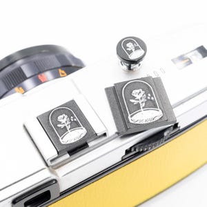 以下が含まれることがあります： 黄色の革製グリップが付いたシルバーのカメラには、ガラスドームの下にバラのデザインが施された3つの黒いアクセサリーがあります。アクセサリーはボタンと2つの長方形のアタッチメントです。バラのデザインは白です。