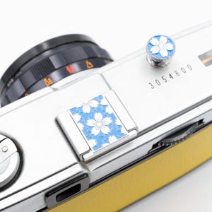 以下が含まれることがあります： 黄色い革のグリップが付いたシルバーのカメラ。フラッシュシューとシャッターボタンには、青と白の花柄のデザインが施されています。レンズは黒色で、カメラ本体には「3054800」の数字が印刷されています。