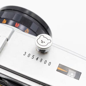 以下が含まれることがあります： 黒いレンズと白い背景の銀色のカメラのクローズアップ。 カメラには、クマの顔のデザインの小さな丸いボタンが付いています。 番号「3054800」がカメラ本体に印刷されています。