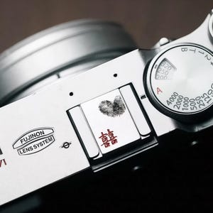 以下が含まれることがあります： 黒いレンズシステムを備えた銀色のカメラのクローズアップ。 カメラには数字と文字のダイヤル、指紋のデザインと漢字が施された小さな長方形のプレートがあります。 「FUJINON LENS SYSTEM」の文字も表示されています。